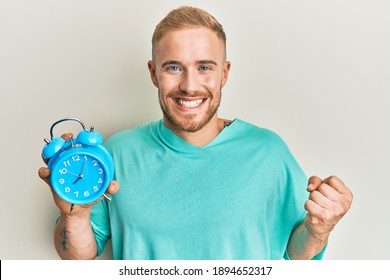 Young caucasian man holding alarm clock screaming proud, celebrating victory and success very excited with raised arm 