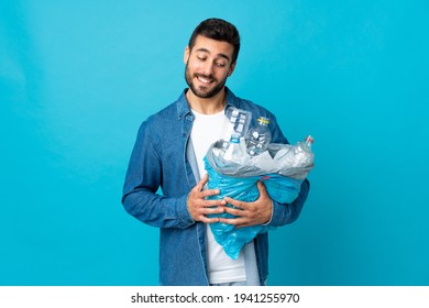 Young caucasian man holding a bag full of plastic bottles to recycle isolated on blue background with happy expression