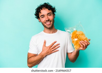 Young Caucasian Man Holding A Bag Of Chips Isolated On Blue Background Laughs Out Loudly Keeping Hand On Chest.