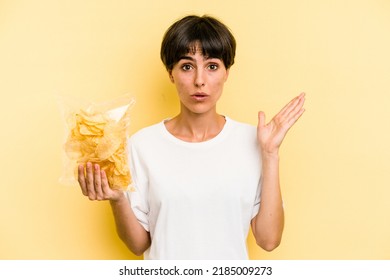 Young caucasian man holding crisps isolated on yellow background surprised and shocked.