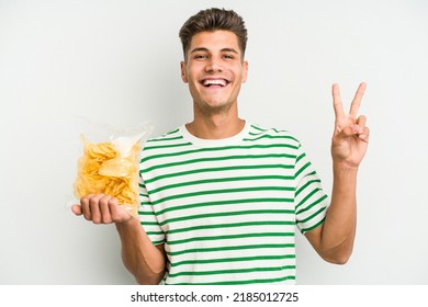 Young caucasian man holding crisps isolated on white background joyful and carefree showing a peace symbol with fingers.