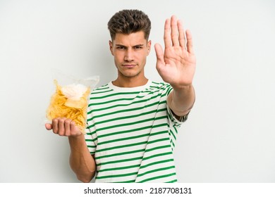 Young caucasian man holding crisps isolated on white background standing with outstretched hand showing stop sign, preventing you.
