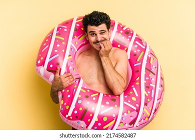 Young caucasian man holding an inflatable donut isolated on yellow background biting fingernails, nervous and very anxious.