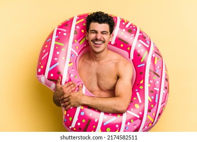 Young caucasian man holding an inflatable donut isolated on yellow background laughing and having fun.