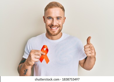 Young Caucasian Man Holding Orange Ribbon For Leukemia Awareness Smiling Happy And Positive, Thumb Up Doing Excellent And Approval Sign 