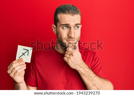 Young caucasian man holding paper with sagittarius zodiac sign serious face thinking about question with hand on chin, thoughtful about confusing idea 