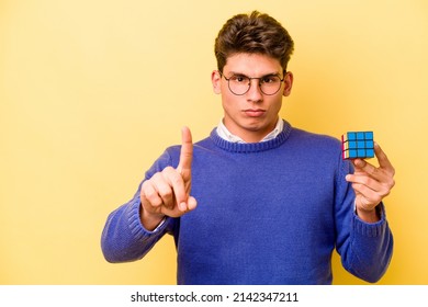 Young Caucasian Man Holding A Rubiks Cube Isolated On Yellow Background Showing Number One With Finger.