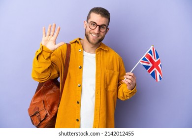 Young caucasian man holding an United Kingdom flag isolated on purple background saluting with hand with happy expression