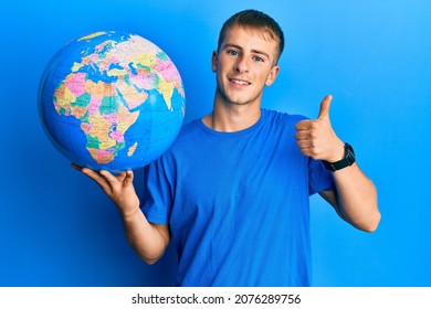 Young caucasian man holding world ball smiling happy and positive, thumb up doing excellent and approval sign 