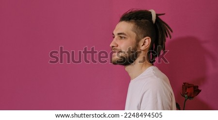 Young Caucasian man holds one red rose behind his back and smiles, wants to give flower. Isolated on pink background. Web banner. Concept of Valentine's Day. International Women's Day greeting card.