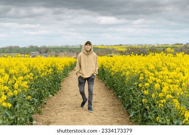 A young Caucasian man in a hoodie walking on a pathway through the white mustard field - Powered by Shutterstock