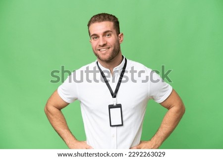Young caucasian man with ID card isolated on green chroma background posing with arms at hip and smiling
