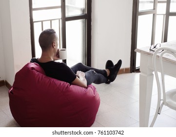 A Young Caucasian Man Indoors Have A Coffee Sitting In A Comfortable Red Bean Bag Chair