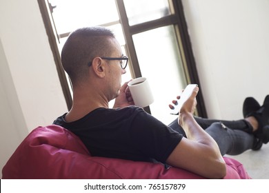 A Young Caucasian Man Indoors Have A Coffee While Is Using His Smartphone Sitting In A Comfortable Red Bean Bag Chair