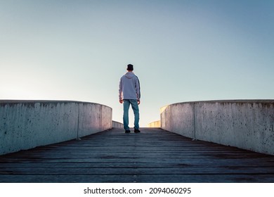 Young Caucasian Man In Jeans, Hat And Hoodie Standing On Concrete Bridge. Rear View Male Pose In City Environment And Clear Sky