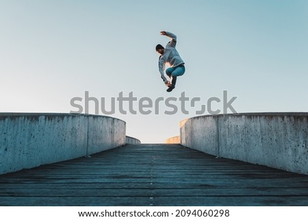 Young caucasian man in jeans and hoodie jumping on concrete bridge. Mid air parkour pose in city environment and clear sky