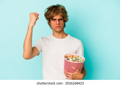 Young caucasian man with makeup holding popcorn isolated on blue background  showing fist to camera, aggressive facial expression. - Powered by Shutterstock