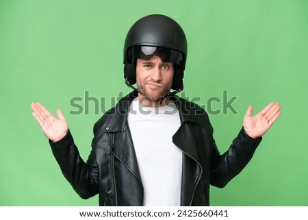 Young caucasian man with a motorcycle helmet isolated on green chroma background having doubts while raising hands