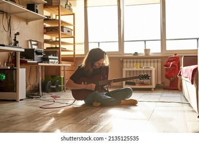 Young caucasian man playing electric guitar notes and chords on floor at sunny home. Domestic hobby, entertainment and leisure. Focused professional male rock musician and songwriter wearing earphones