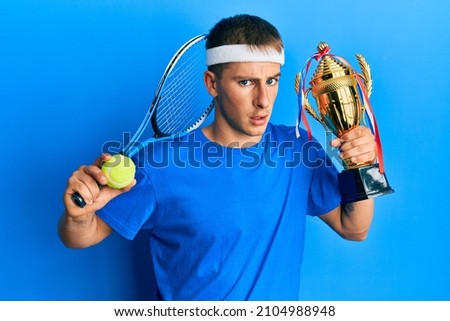 Young caucasian man playing tennis holding trophy clueless and confused expression. doubt concept. 