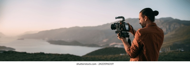 A young Caucasian man with a professional camera in his hands on a mountain by the sea.  The operator holds a video camera, shoots a sunset landscape, mountains, sea, setting sun - Powered by Shutterstock