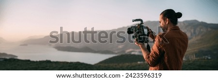 A young Caucasian man with a professional camera in his hands on a mountain by the sea.  The operator holds a video camera, shoots a sunset landscape, mountains, sea, setting sun
