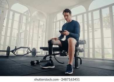 Young caucasian man with prosthetic leg sitting using mobile phone on bench press at the gym - Powered by Shutterstock