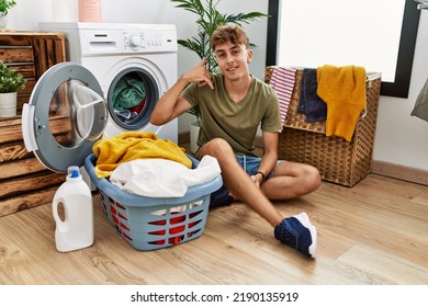 Young caucasian man putting dirty laundry into washing machine smiling doing phone gesture with hand and fingers like talking on the telephone. communicating concepts.  - Shutterstock ID 2190135919