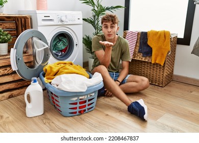 Young Caucasian Man Putting Dirty Laundry Into Washing Machine Looking At The Camera Blowing A Kiss With Hand On Air Being Lovely And Sexy. Love Expression. 