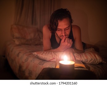 Young Caucasian Man Reads A Book In Bed By Candlelight