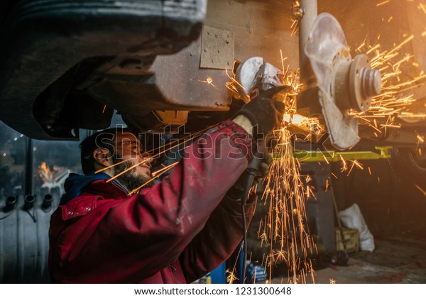Young caucasian man in red\
work clothing repairing car with angle cutter. Many sparks on\
background.