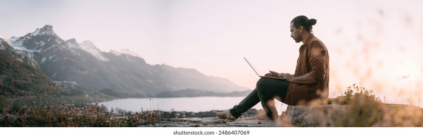 A young Caucasian man remotely works with a laptop in a garden on a mountain overlooking the sea and sunset.  The guy is sitting on a rock with a laptop on his lap in the rays of the sun. - Powered by Shutterstock