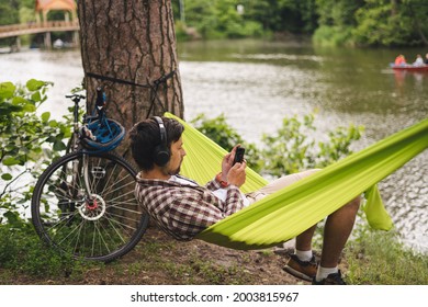 Young caucasian man resting in hammock, listening to music on headphones and using smartphone afterwards on bicycle in forest near the lake. Audio healing. Idyllic place. Travel, camping in nature. - Powered by Shutterstock