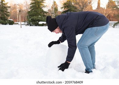 Young Caucasian Man Rolling A Big Snowball To Make Snowman