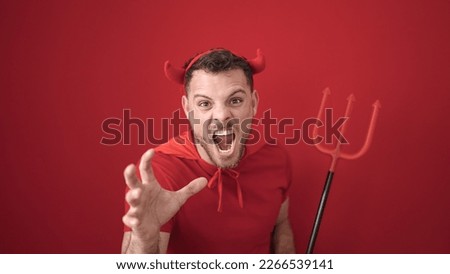 Young caucasian man screaming wearing devil costume over isolated red background