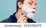 Young Caucasian man shaving, applying shaving cream, using a razor. Close-up of a man shaving, focusing on shaving cream and razor.