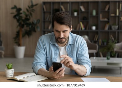 Young Caucasian Man Sit At Desk At Home Look At Cellphone Screen Texting Or Messaging Online On Gadget. Millennial Male User Or Client Use Modern Smartphone, Browse Internet. Technology Concept.