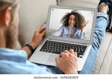 Young caucasian man sit on sofa video calling remote friend african woman talking in online webcam chat on laptop screen at home office. Distance dating videocall concept. Over shoulder closeup view