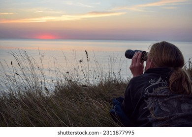 Young caucasian man sits on a cliff and use binoculars for bird watching at the ocean sky, sunset in the baltic sea coast while seagulls and swallows fly around. Solo summer holiday at the beach