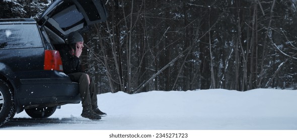 Young Caucasian man sits in trunk of car and calls on phone mechanic for help. broken-down vehicle in snowy winter forest. empty rural road. Horizontal web banner with copy space, side view - Powered by Shutterstock