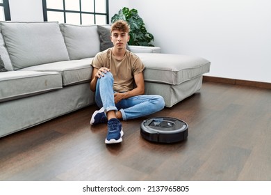 Young Caucasian Man Sitting At Home By Vacuum Robot Thinking Attitude And Sober Expression Looking Self Confident 