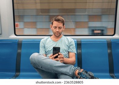 Young Caucasian man sitting in the train on the subway and using mobile phone. Male traveler in metro train surfing internet, checking social media on smartphone