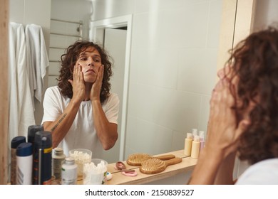 Young Caucasian Man Smearing After Shave Balm On His Face And Looking At Himself In Mirror In Bathroom. Curly Guy With Tattoo Wearing T-shirt. Modern Domestic Lifestyle. Hygiene And Face Skin Care