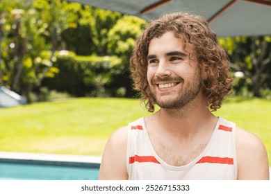 A young Caucasian man smiles warmly near a pool outdoors with copy space at home. He has curly brown hair, light stubble, and is wearing a casual sleeveless top, unaltered. - Powered by Shutterstock