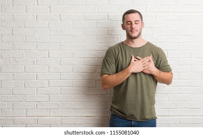 Young caucasian man standing over white brick wall smiling with hands on chest with closed eyes and grateful gesture on face. Health concept.