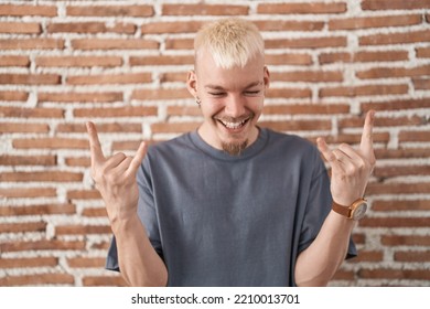 Young Caucasian Man Standing Over Bricks Wall Shouting With Expression Doing Rock Symbol With Hands Up. Music Star. Heavy Music Concept. 