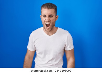 Young caucasian man standing over blue background angry and mad screaming frustrated and furious, shouting with anger. rage and aggressive concept.  - Shutterstock ID 2349198099