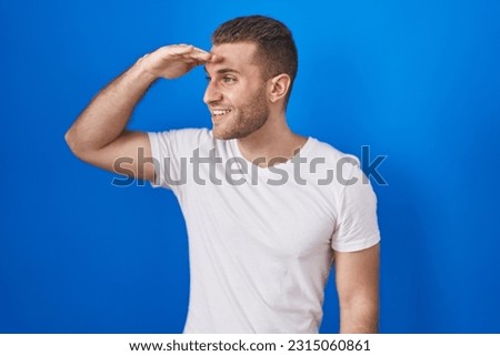 Young caucasian man standing over blue background very happy and smiling looking far away with hand over head. searching concept. 