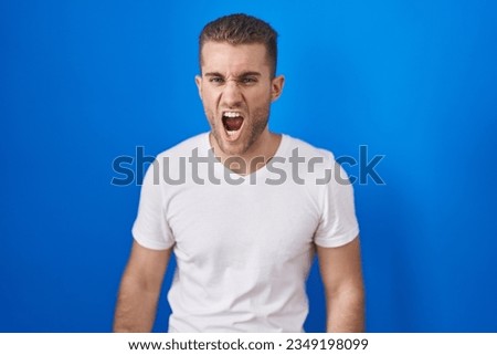 Young caucasian man standing over blue background angry and mad screaming frustrated and furious, shouting with anger. rage and aggressive concept. 