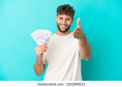 Young Caucasian Man Taking A Lot Of Money Isolated On Blue Background Shaking Hands For Closing A Good Deal